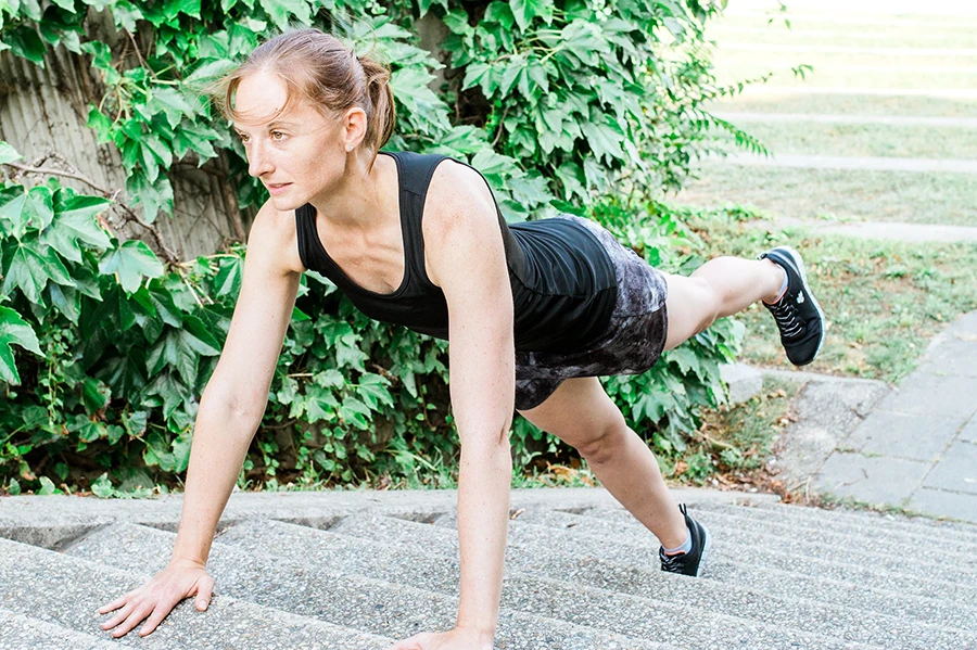 Ursula führt eine Plank auf einer Treppen im Park aus. Sie hebt ihre Beine abwechselnd an.